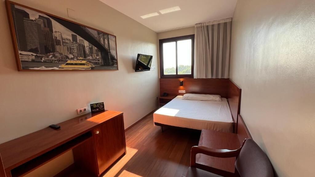 A hotel room with a bed, bedside table, desk, chair, TV, and a large cityscape painting on the wall.