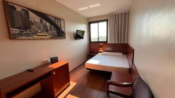 A hotel room with a bed, bedside table, desk, chair, TV, and a large cityscape painting on the wall.