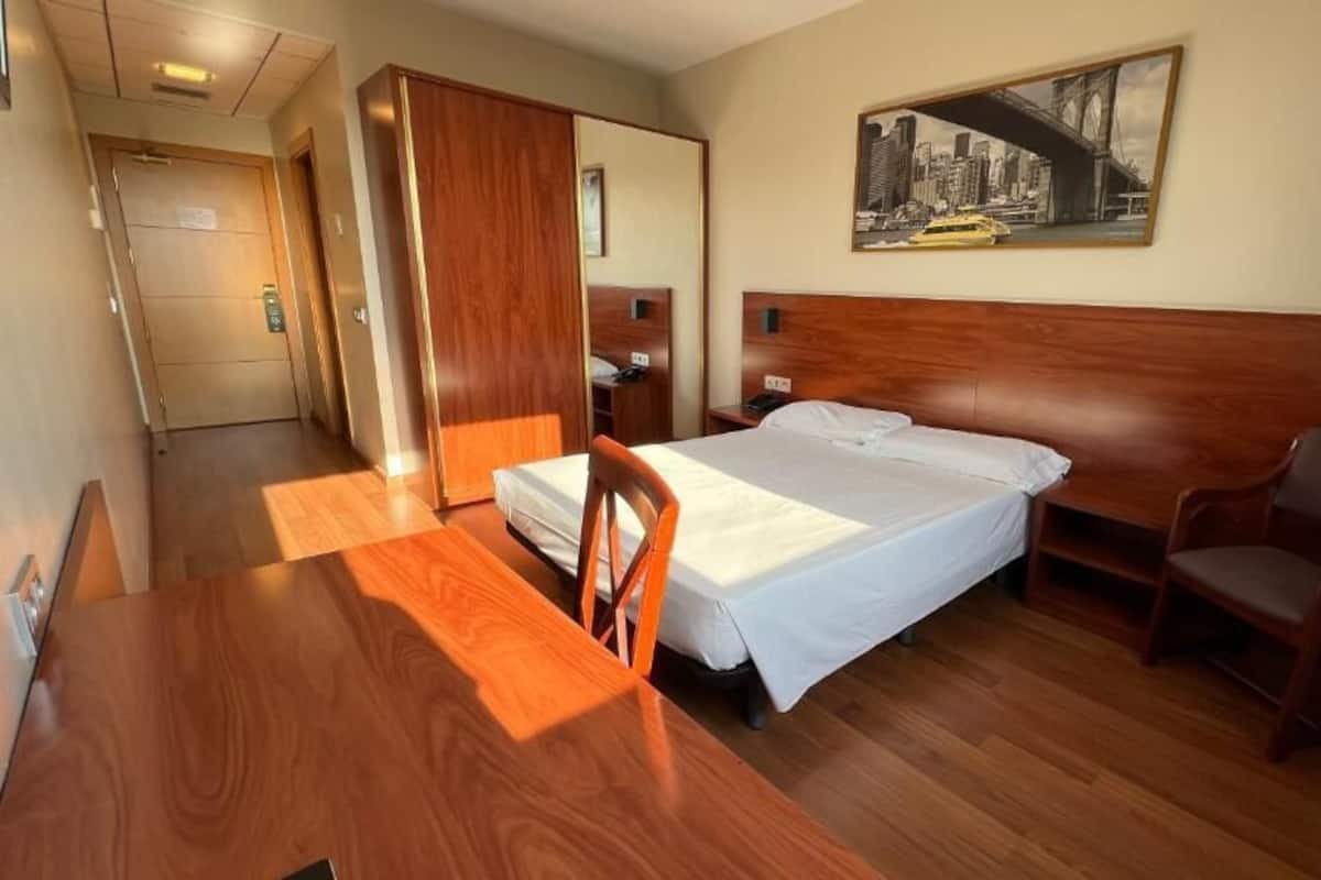 A hotel room with a bed, a wooden desk, a chair, a nightstand, and a painting on the wall.