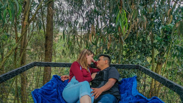 A couple sitting on a hammock in a natural setting with a wire fence and greenery in the background. - Samay Collections (Tababela)