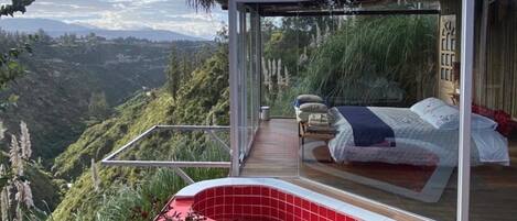 A modern bedroom with a glass enclosure, a red hot tub, and a bed with a view of a mountainous landscape.