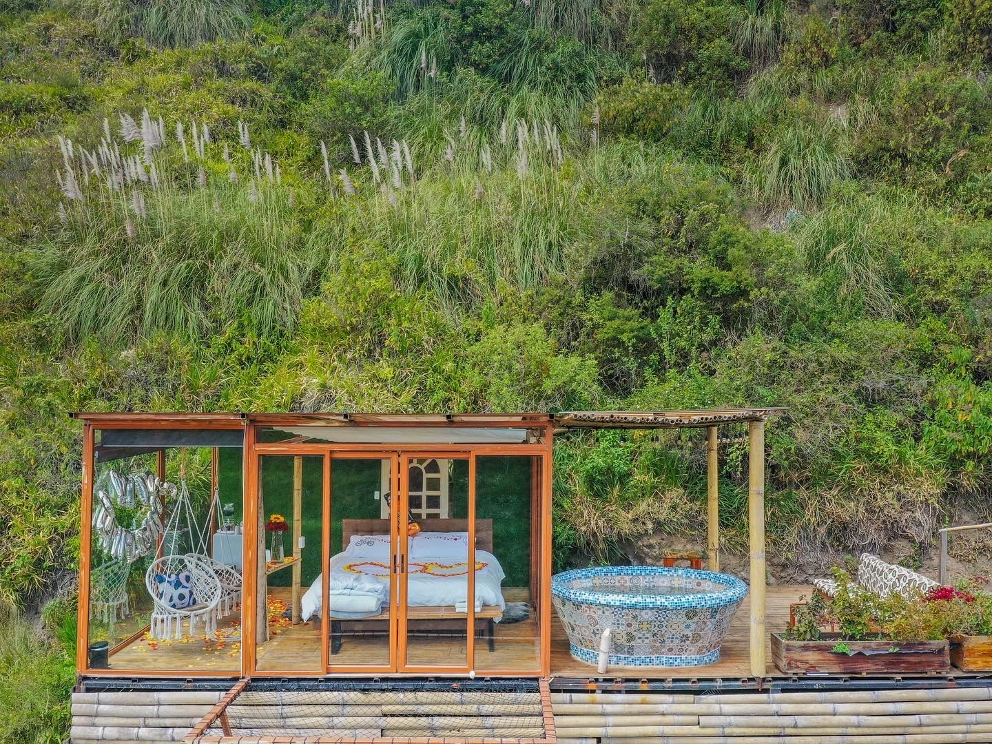 A glass-enclosed room with a bed, a hanging chair, and a mosaic hot tub.