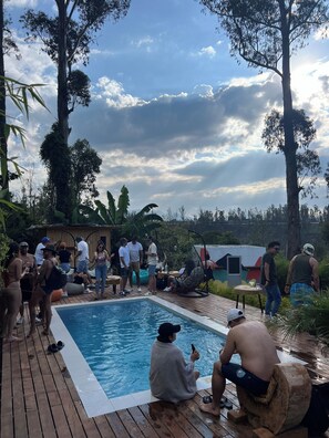 Une terrasse en bois avec une piscine, entourée d’arbres et d’une petite construction en bois.