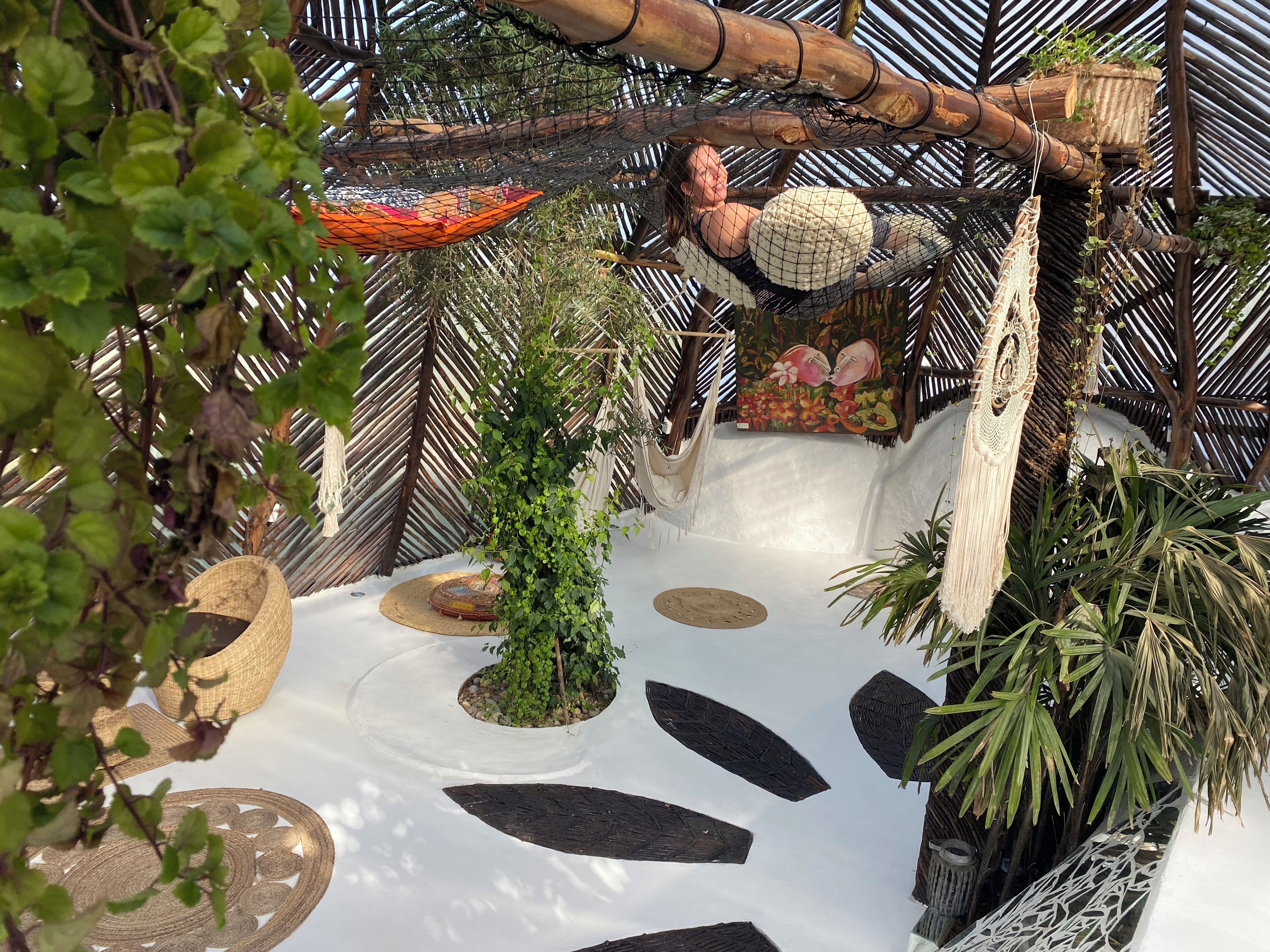 A hammock suspended from a bamboo structure, surrounded by lush greenery and a decorative painting.