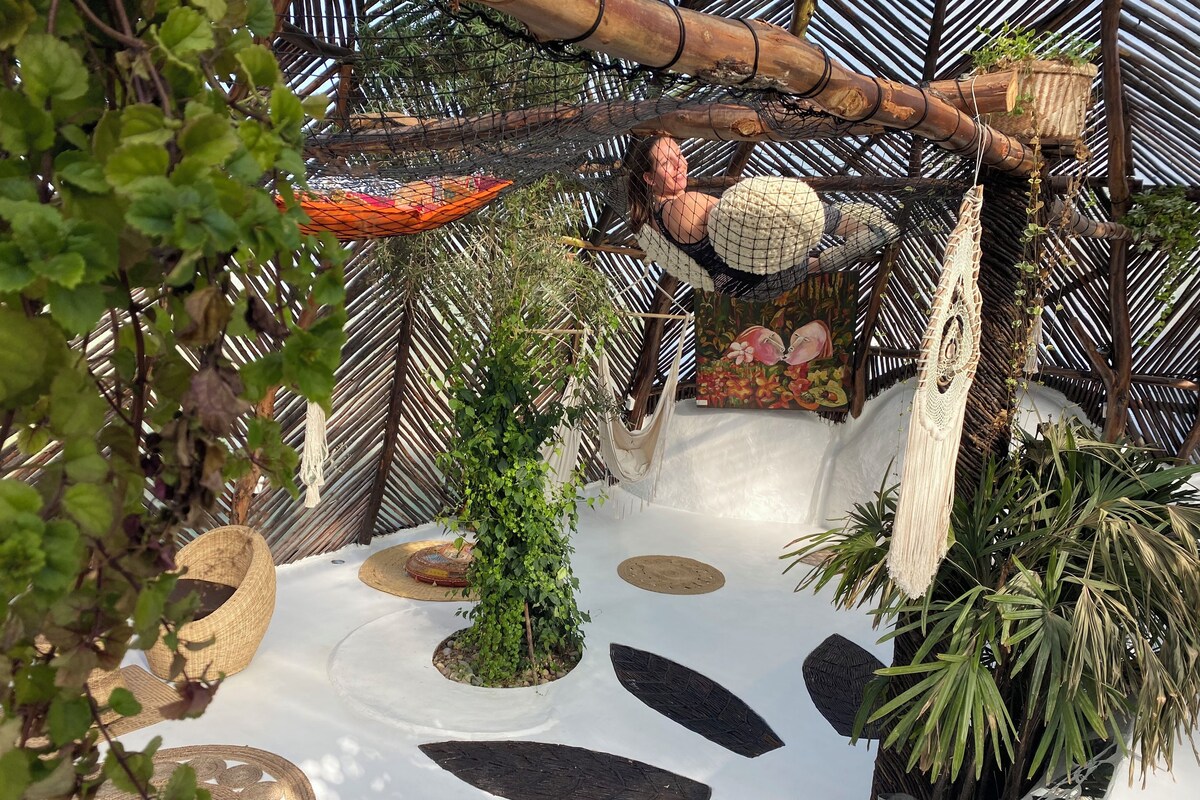 A hammock suspended from a bamboo structure, surrounded by lush greenery and a decorative painting.