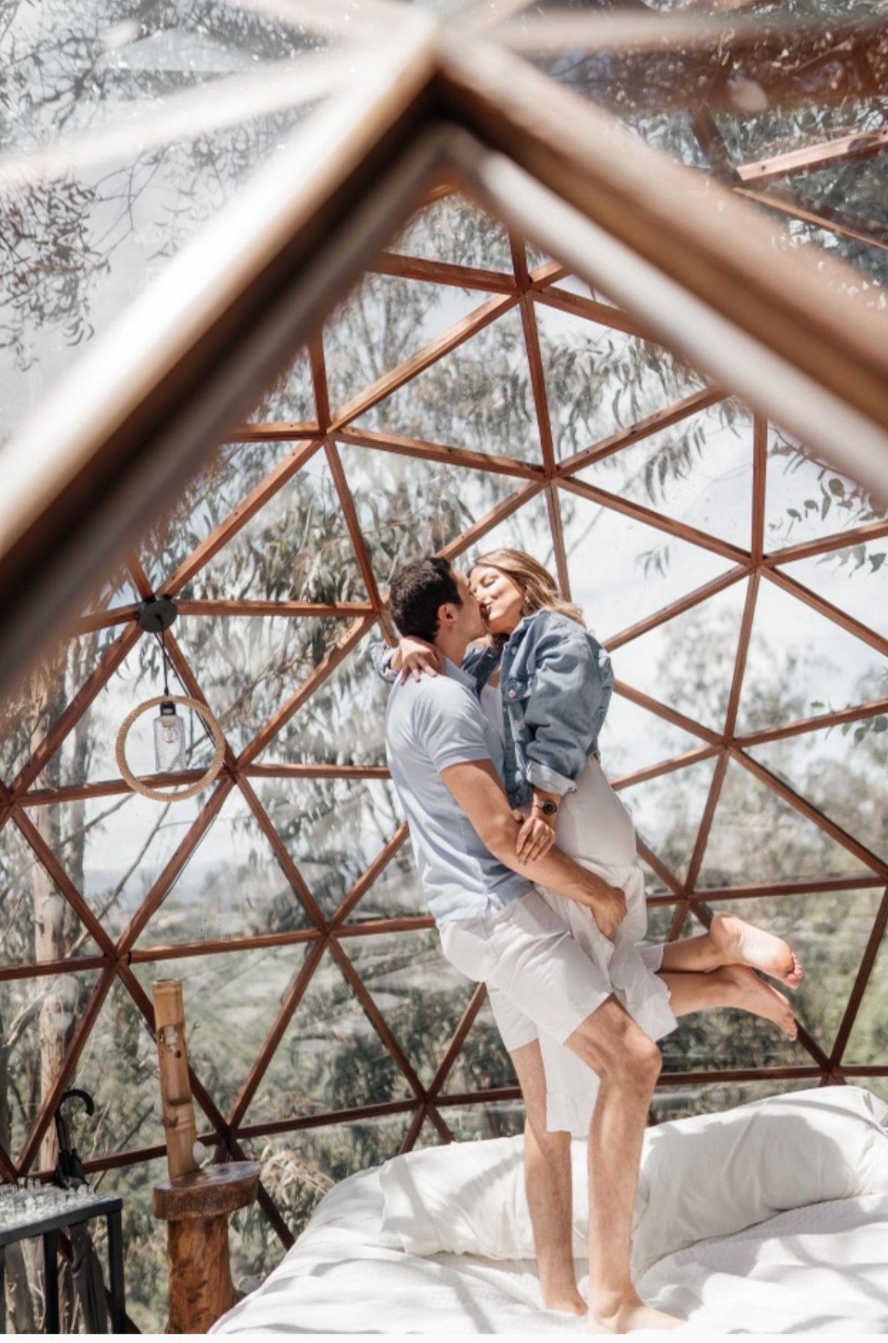 A couple in a geodesic dome structure with a bed, a hanging light fixture, and a bamboo chair.