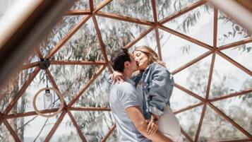 A couple in a geodesic dome structure with a bed, a hanging light fixture, and a bamboo chair.