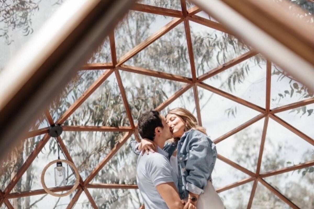 A couple in a geodesic dome structure with a bed, a hanging light fixture, and a bamboo chair.