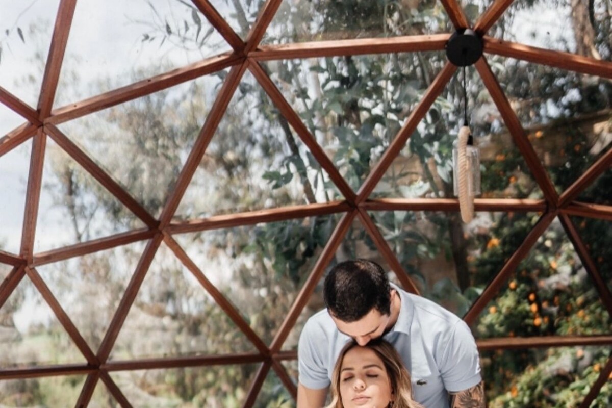 A geometric metal structure with a person sitting on a white bed, another person standing behind them, and a view of greenery outside.