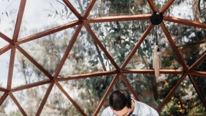 A geometric metal structure with a person sitting on a white bed, another person standing behind them, and a view of greenery outside. - Samay Collections (Tababela)