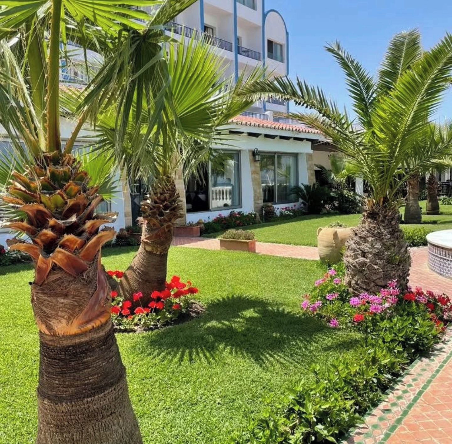 A well-maintained garden with palm trees, red flowers, and a building with a tiled roof.