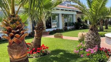 A well-maintained garden with palm trees, red flowers, and a building with a tiled roof.