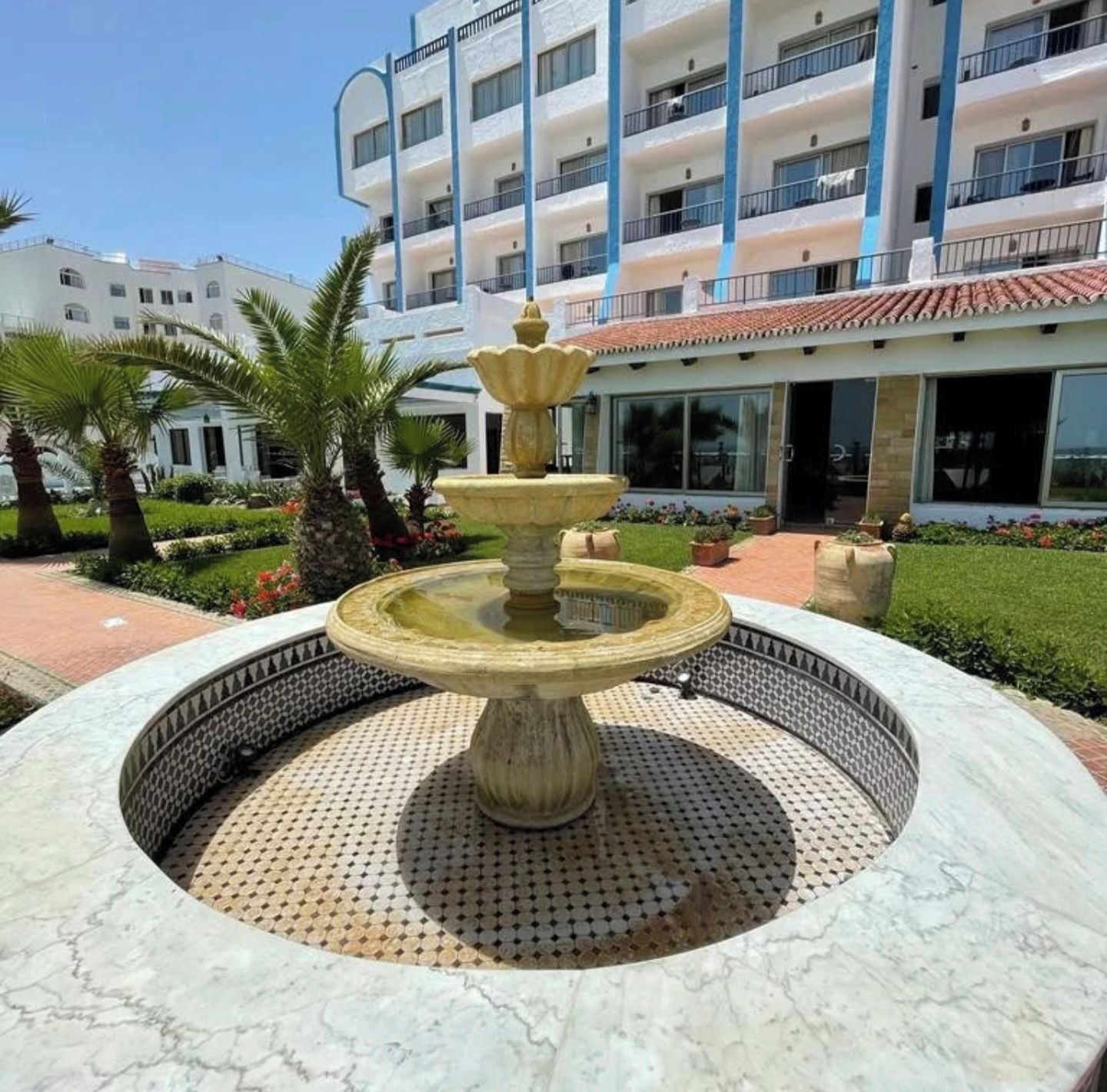 A fountain with a tiered design in a circular stone basin, surrounded by a landscaped garden with palm trees and a building with multiple balconies in the background.