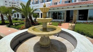 Uma fonte com design em níveis numa bacia de pedra circular, rodeada por um jardim ajardinado com palmeiras e um edifício com varandas múltiplas ao fundo.