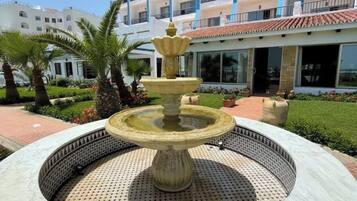 A fountain with a tiered design in a circular stone basin, surrounded by a landscaped garden with palm trees and a building with multiple balconies in the background.
