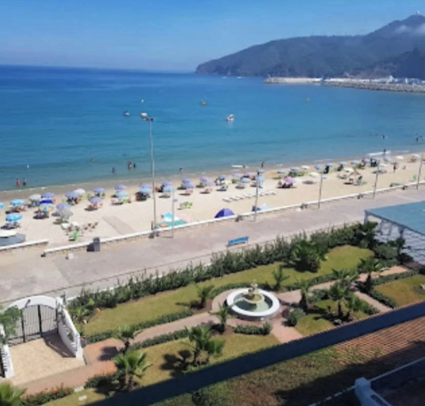 Een strand met parasols, mensen, een fontein en een berg op de achtergrond.