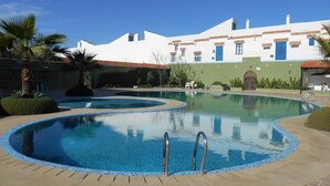 Piscina com design curvo, cercada por um muro baixo e área de pátio. Palmeiras e um edifício com fachada branca e detalhes azuis ao fundo.