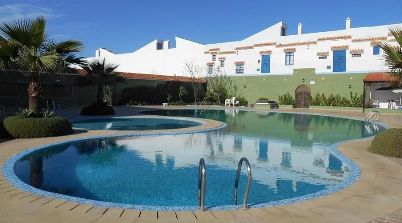 A swimming pool with a curved edge, a metal ladder, and a building with white walls and blue doors in the background.