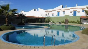 Uma piscina de borda curva, com uma escada de metal, e um edifício com paredes brancas e portas azuis ao fundo.