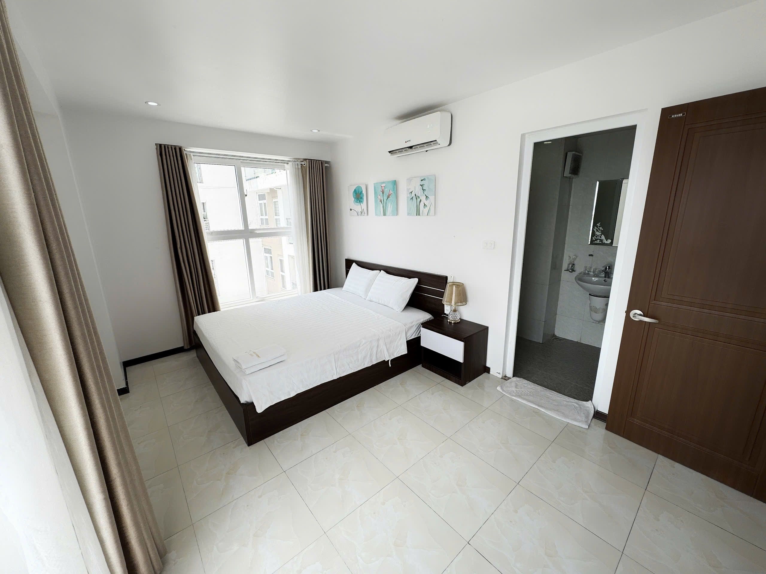 A modern bedroom with a large bed, a bedside table, an air conditioning unit, and a view of the outside through a window.