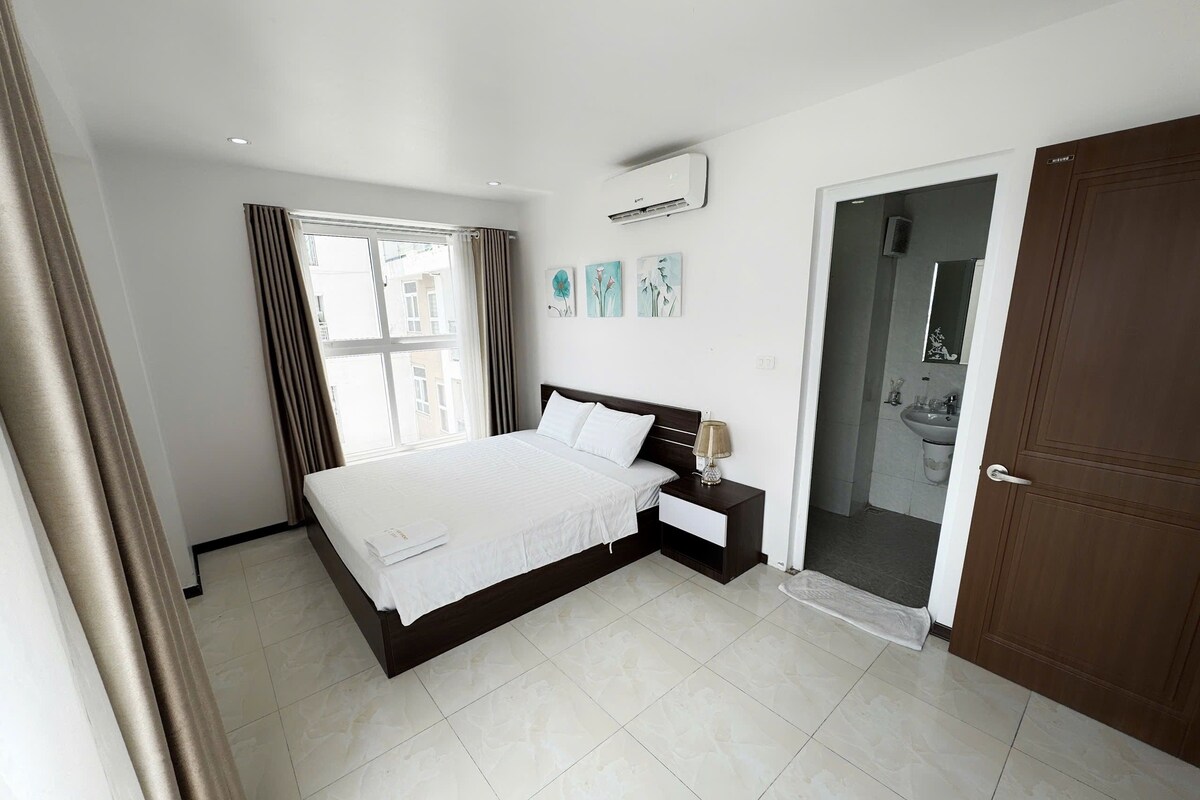 A modern bedroom with a large bed, a bedside table, an air conditioning unit, and a view of the outside through a window.