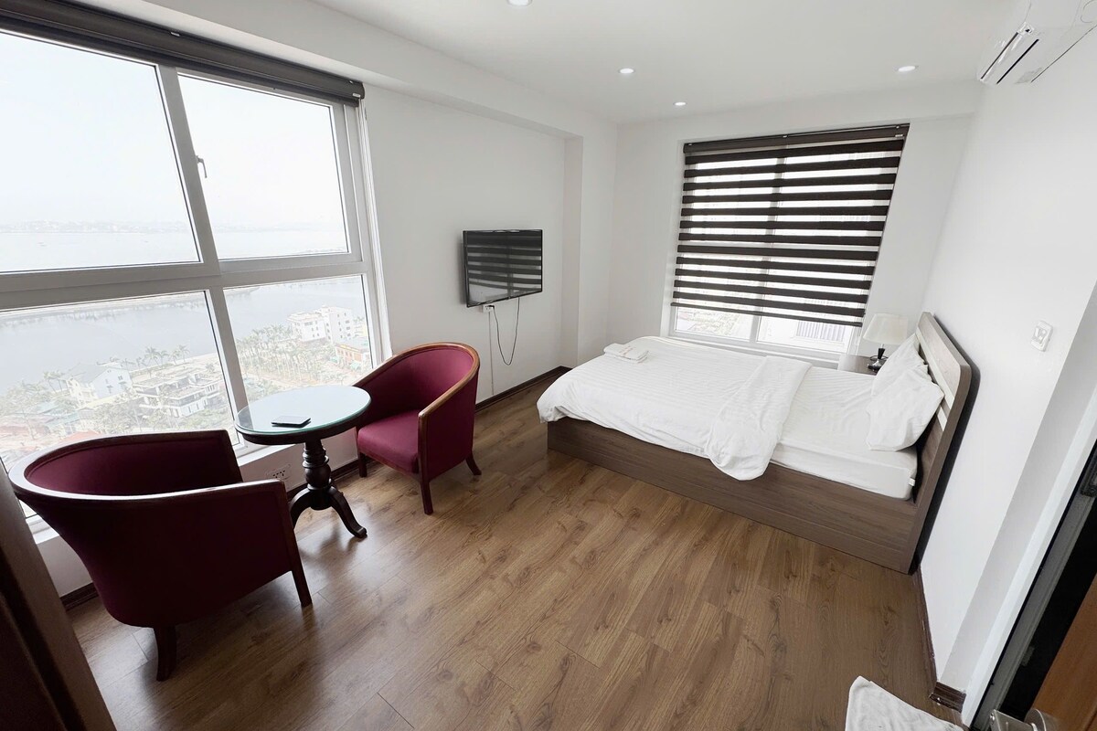 A hotel room with a bed, a red armchair, a small table, a TV, and a large window with blinds.