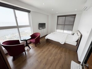 A hotel room with a bed, a red armchair, a small table, a TV, and a large window with blinds. - New Life Tower Bai Chay (Bai Chay)