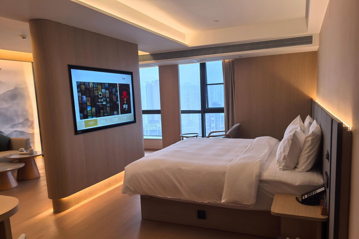 A modern hotel room with a large bed, a desk, a TV mounted on the wall, and a large window with a city view.