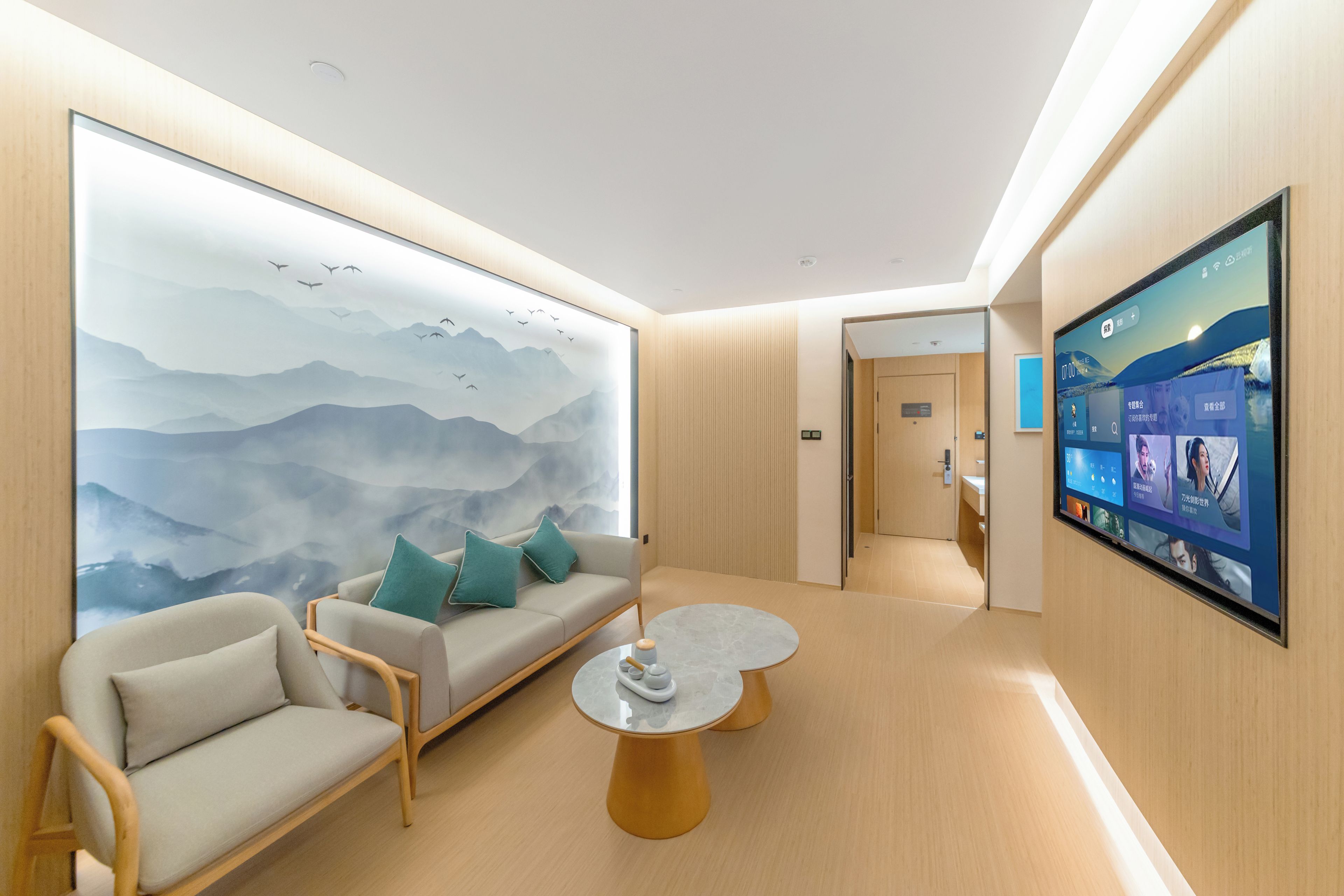 A modern hotel room with a large wall mural, a sofa with cushions, a round coffee table, and a mounted TV screen.
