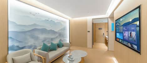 Habitación de hotel moderna con un gran mural en la pared, un sofá con cojines, una mesa de centro redonda y una pantalla de televisión montada.
