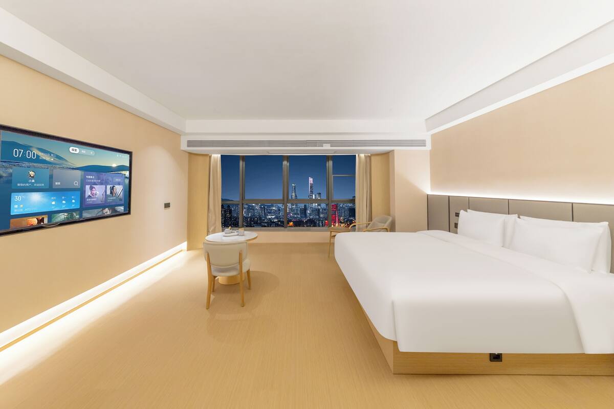 Une chambre d’hôtel avec un grand lit, un bureau, une chaise, une télévision et une vue sur le paysage urbain, visible à travers une grande fenêtre.
