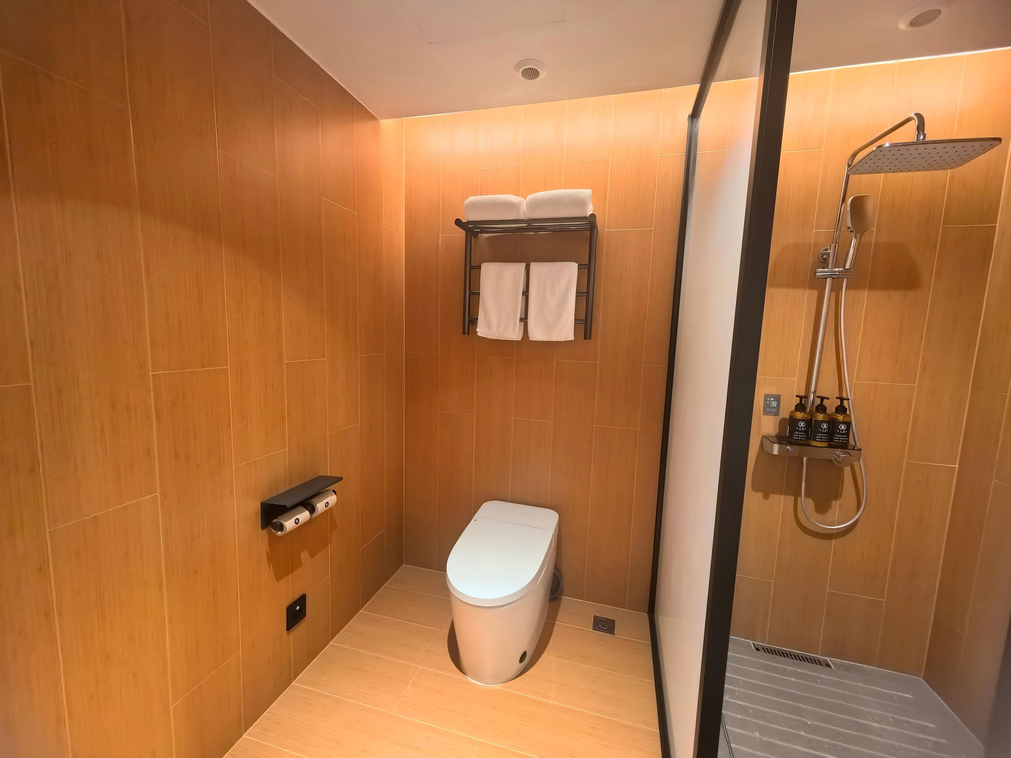 A modern bathroom with wooden walls, a toilet, a shower with a handheld showerhead, and a towel rack with two towels.
