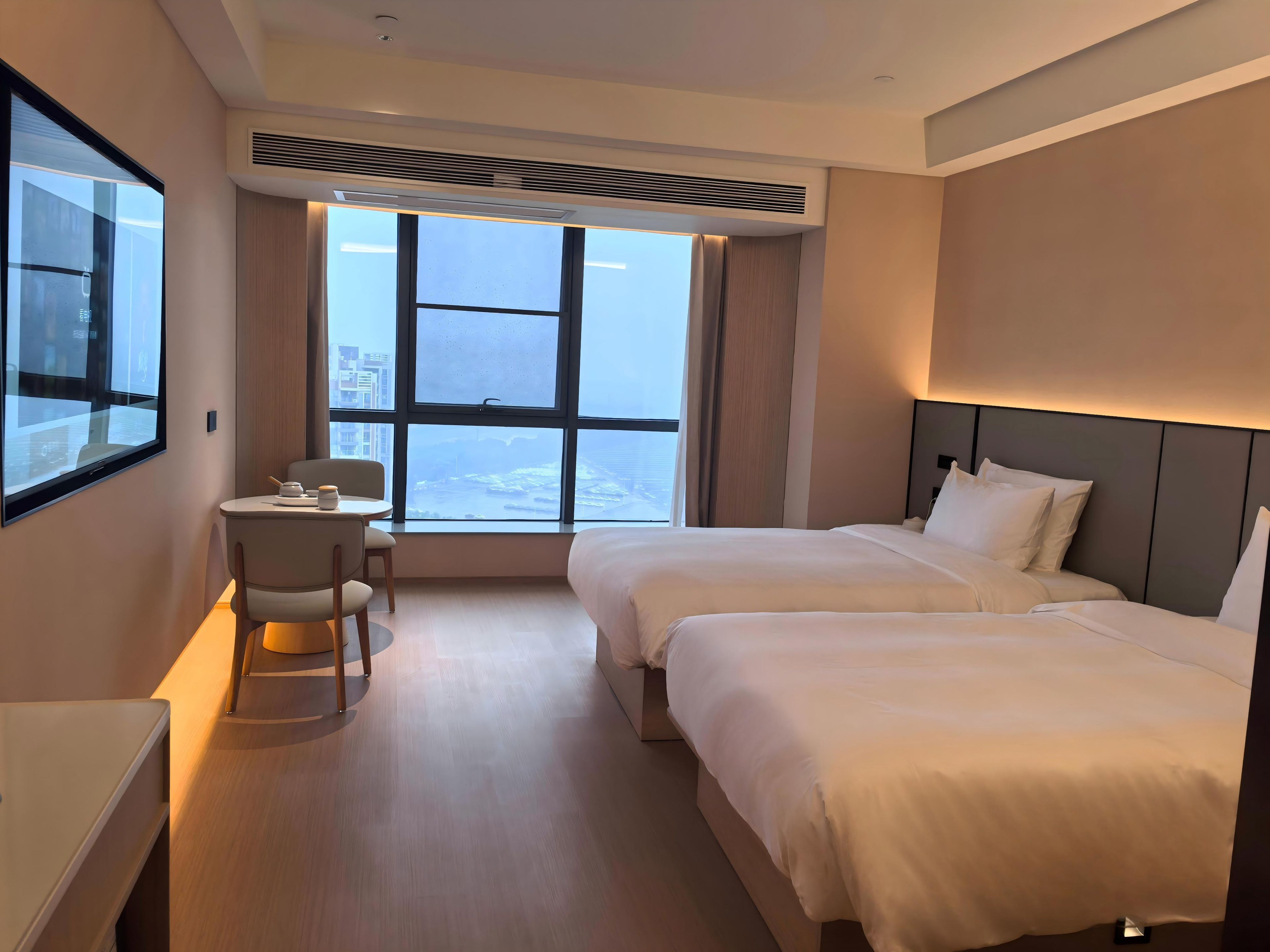 A modern hotel room with a large window, a desk with a chair, and two beds.