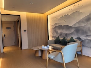 A modern hotel room with a wooden floor, a sofa with green cushions, a round table with a teapot and cups, and a large wall mural of mountains and birds. - Xianze Hotel (Guangzhou)