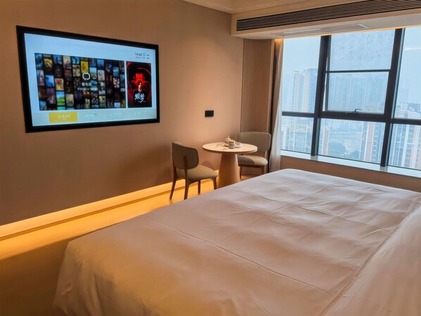 Une chambre d’hôtel avec un grand lit, une petite table ronde avec des chaises, un téléviseur fixé au mur diffusant une émission, et une fenêtre donnant sur la ville.