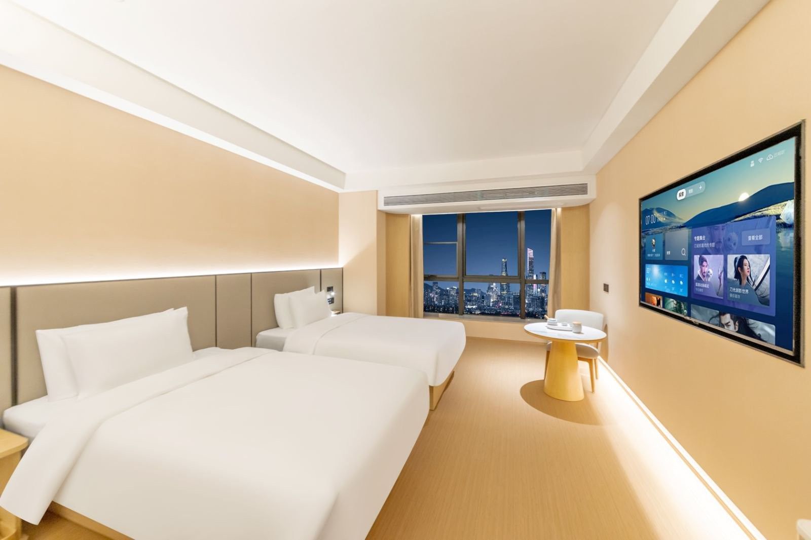 A hotel room with two beds, a small table, and a large TV screen displaying a cityscape view.