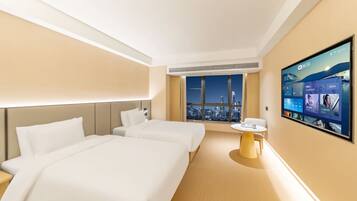 A hotel room with two beds, a small table, and a large TV screen displaying a cityscape view.