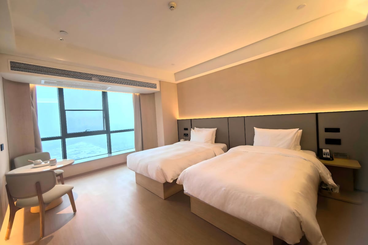 A modern hotel room with two beds, a sitting area, and a large window offering a view.
