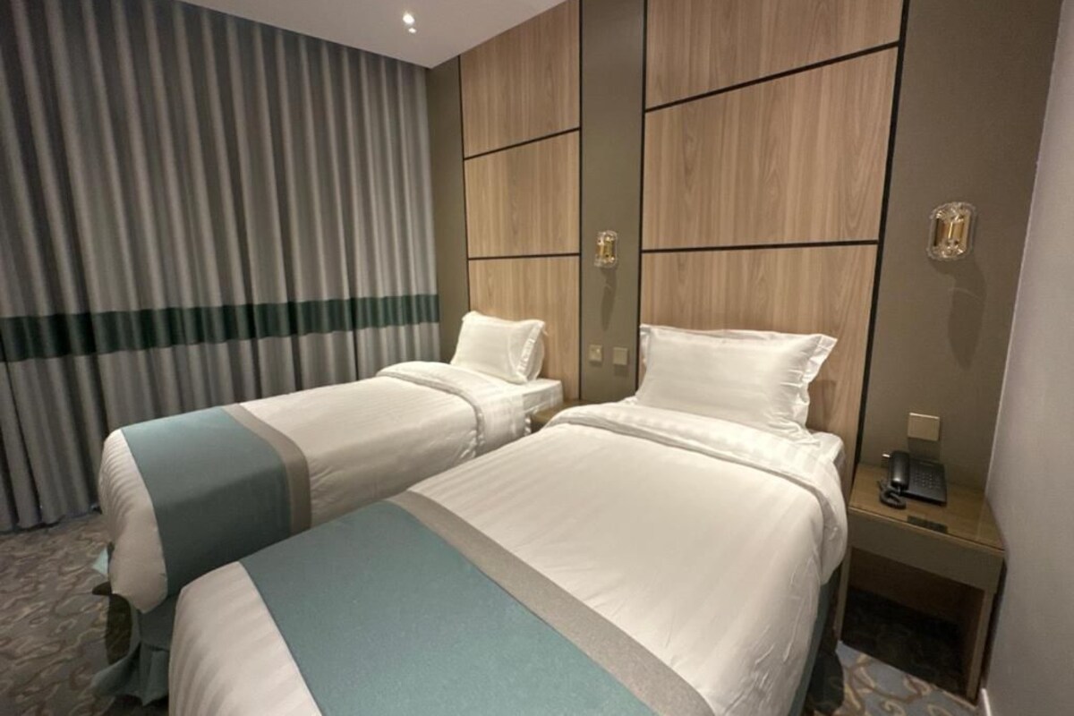 Une chambre d’hôtel avec deux lits, une tête de lit en bois, un bureau avec un téléphone et un rideau.