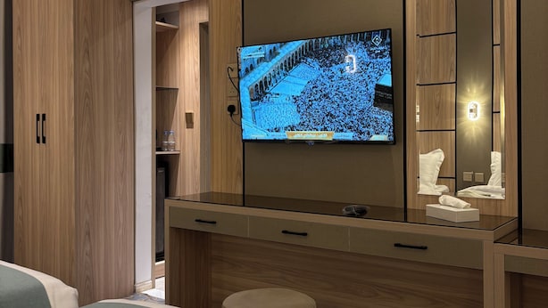 Habitación de hotel con una gran televisión de pantalla plana montada en una pared de madera, una mesita de noche con espejo y una cama con colcha azul y blanca.