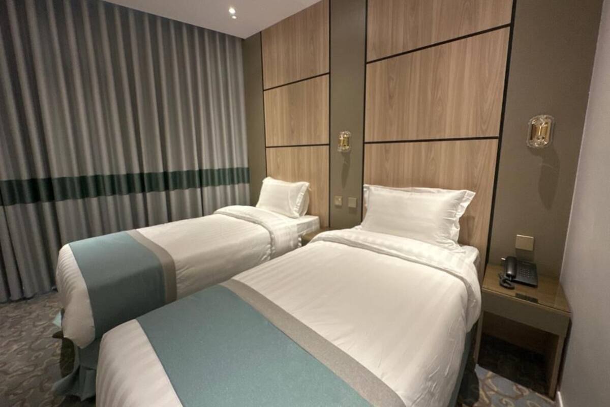 Une chambre d’hôtel avec deux lits, une tête de lit en bois, un bureau avec un téléphone et un rideau.