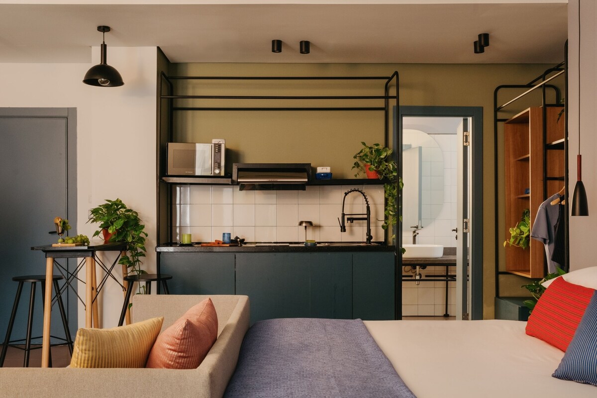 A modern kitchen with a built-in microwave, a sink, and a window. There is a dining area with a table and chairs, and a bed with pillows.