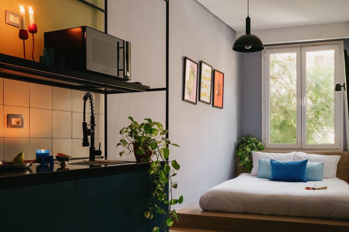 Cocina moderna con microondas, pileta y una planta. Se observa una cama con almohadas y una ventana con vista a follaje.