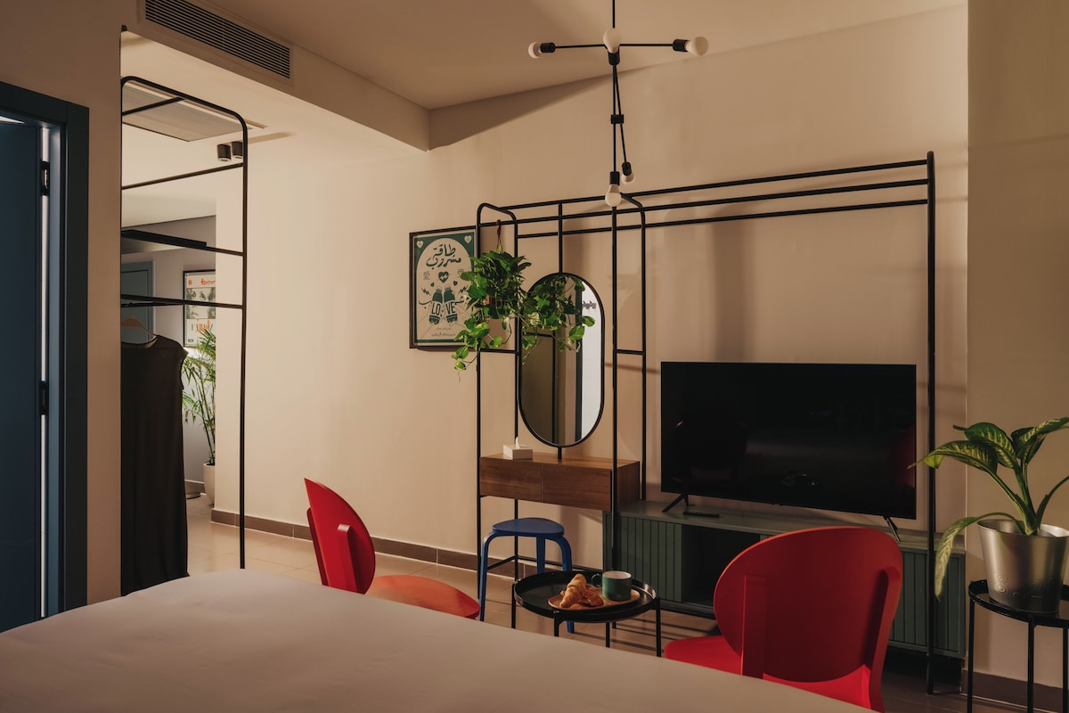 A modern hotel room with a bed, a TV, a desk with a chair, a small table with a cup and a plate, and a potted plant.