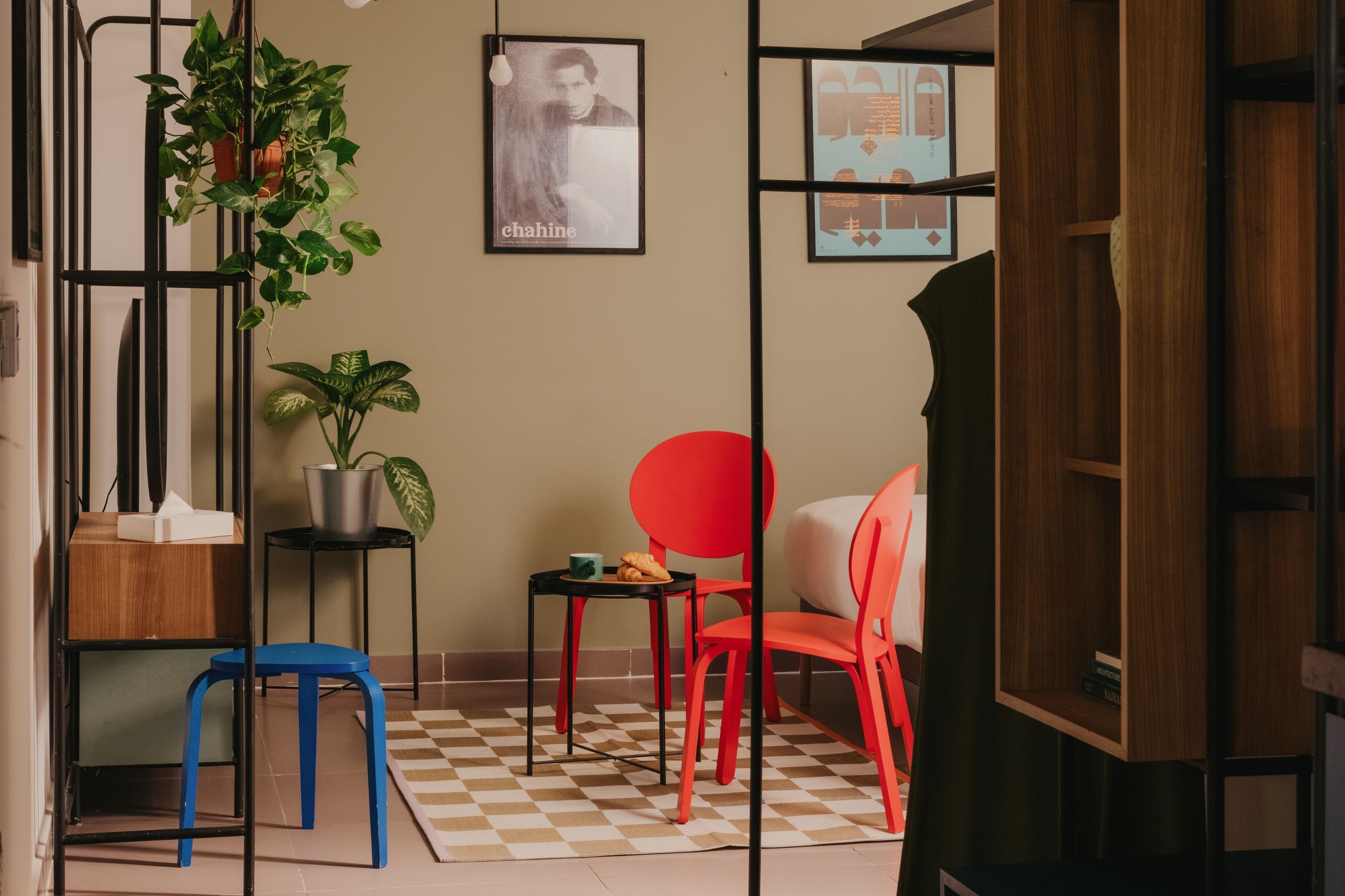 A room with a checkered floor, a blue chair, a red chair, a small table with a cup and a plate, and a plant on a shelf.