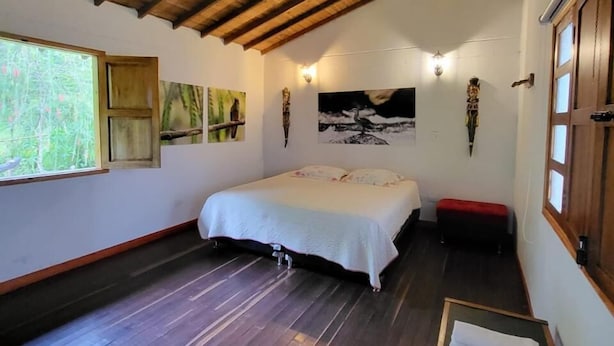 A room with a wooden ceiling, a bed with a white bedspread, a bench, and artwork on the walls. - Kumanday Lodge  (Pereira)