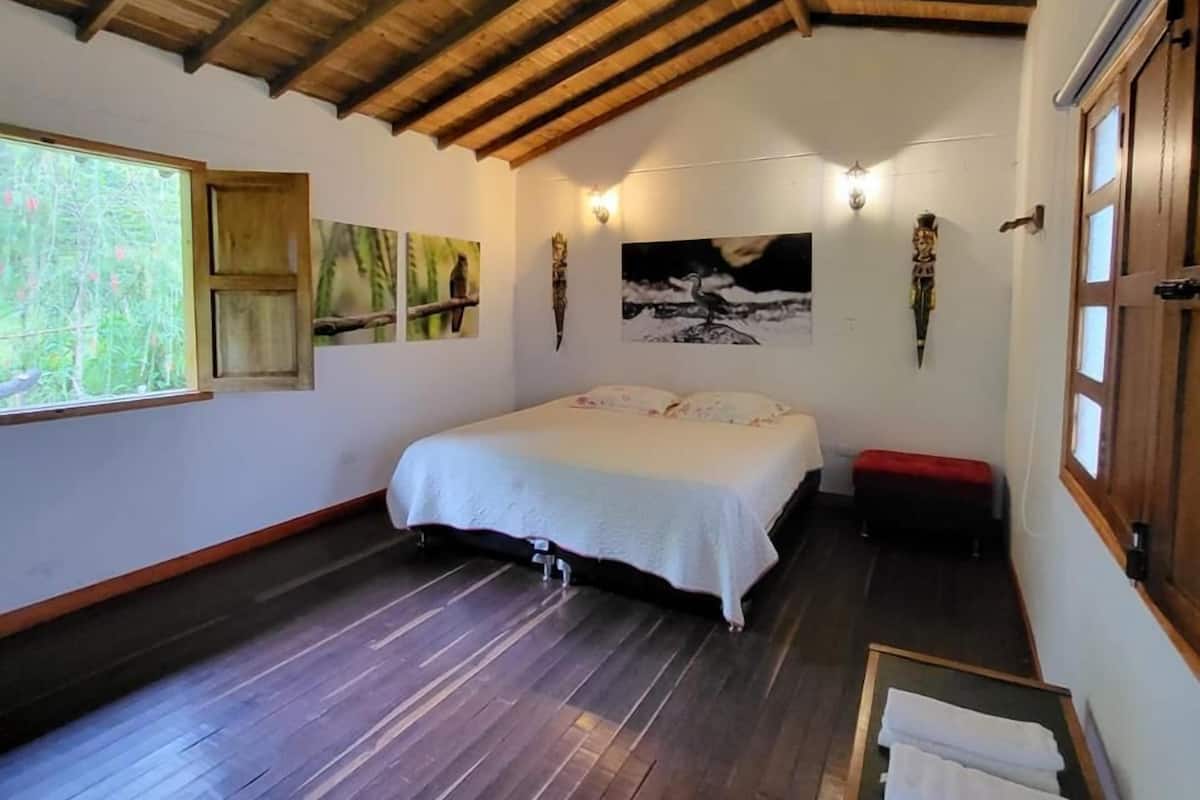 Una habitación con techo de madera, una cama con cubrecama blanco, un banco y obras de arte en las paredes.