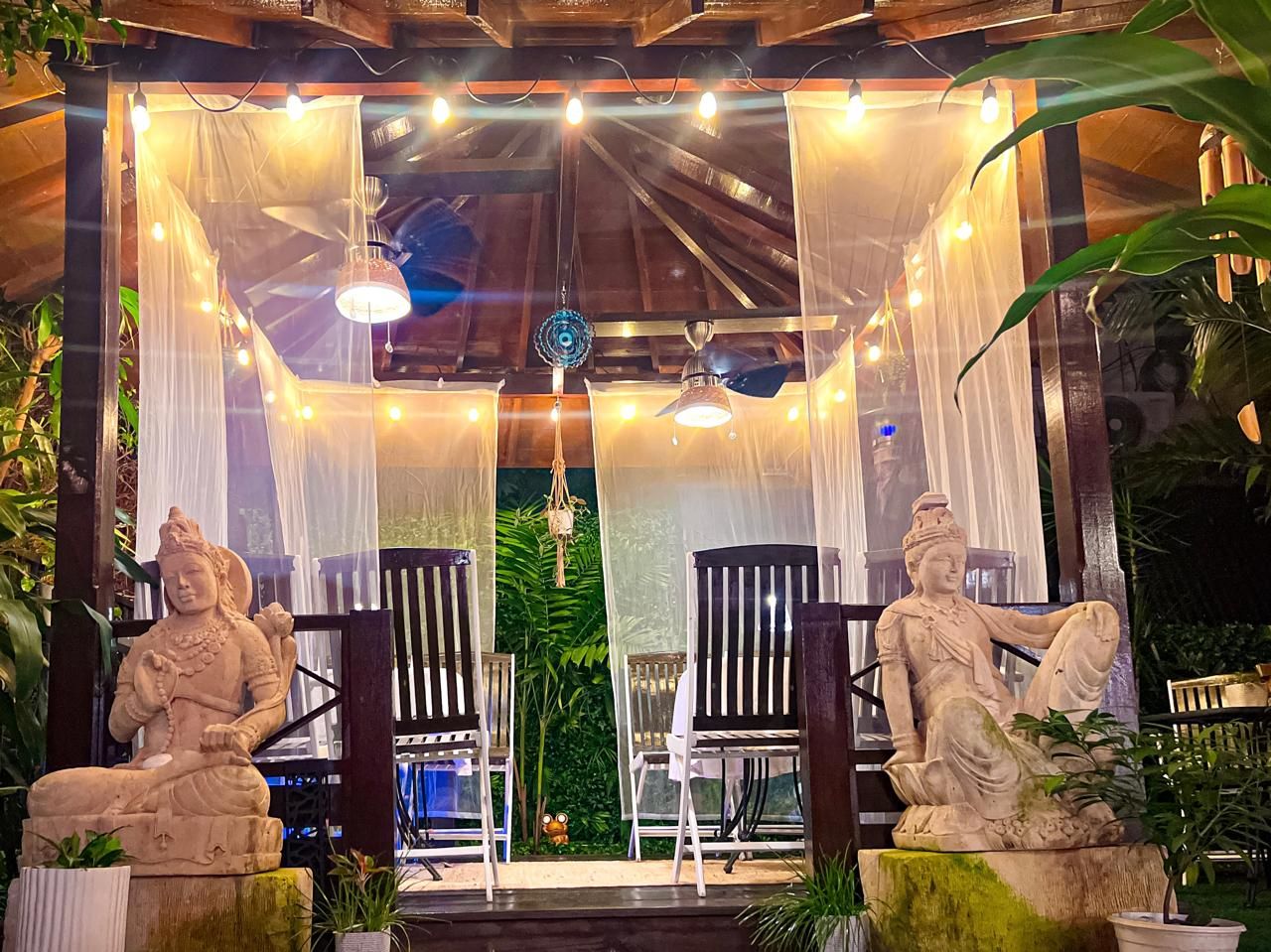 A serene outdoor seating area with two stone statues, wooden chairs, and a canopy.