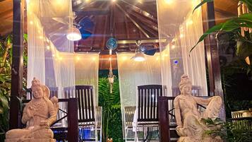 A serene outdoor seating area with two stone statues, wooden chairs, and a canopy.