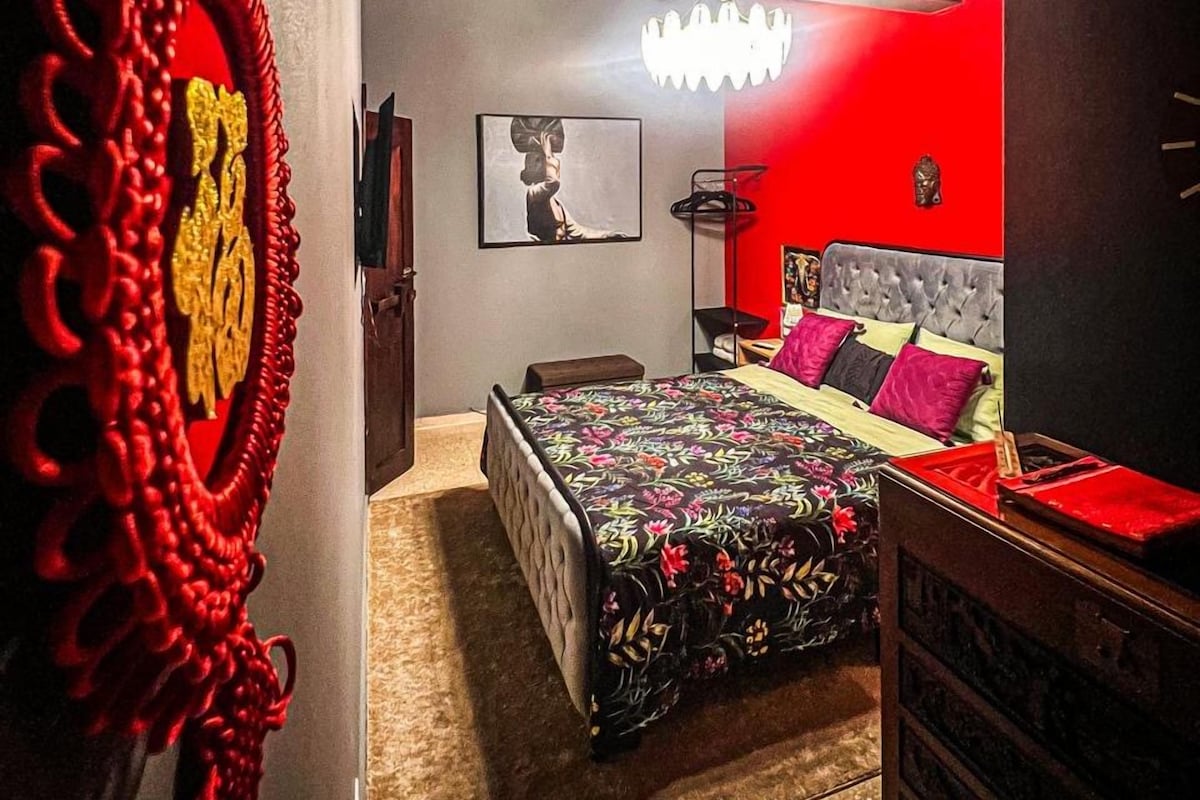 Une chambre à coucher comprenant un lit, une table de chevet, une commode, un mur d’accent rouge et un lustre.